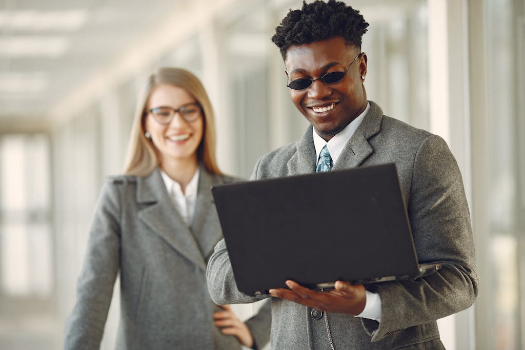Positive smiling stylish diverse business people in grey formal outfits and trendy eyeglasses browsing netbook while standing in contemporary workspace in daylight